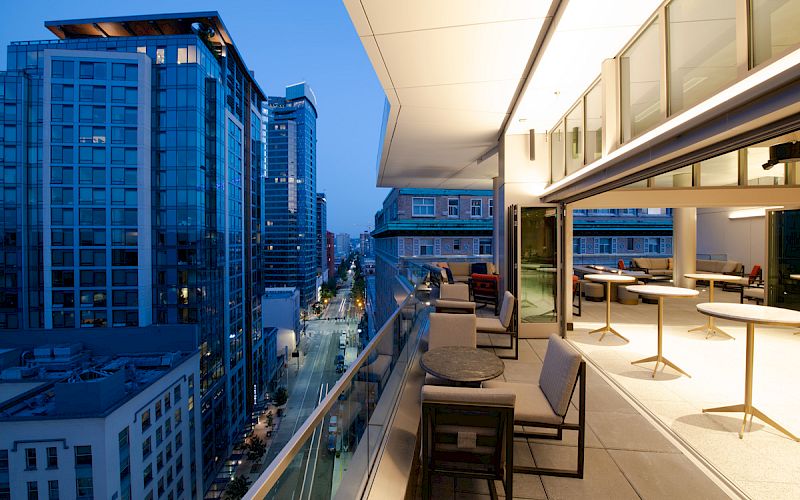 A modern cityscape view from a high-rise balcony at dusk, featuring glass buildings, street lights, and outdoor seating areas.