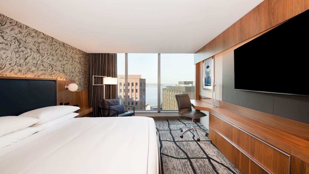 A modern hotel room features a large bed, TV, desk, chair, and a view of buildings through a wide window, with a textured accent wall.