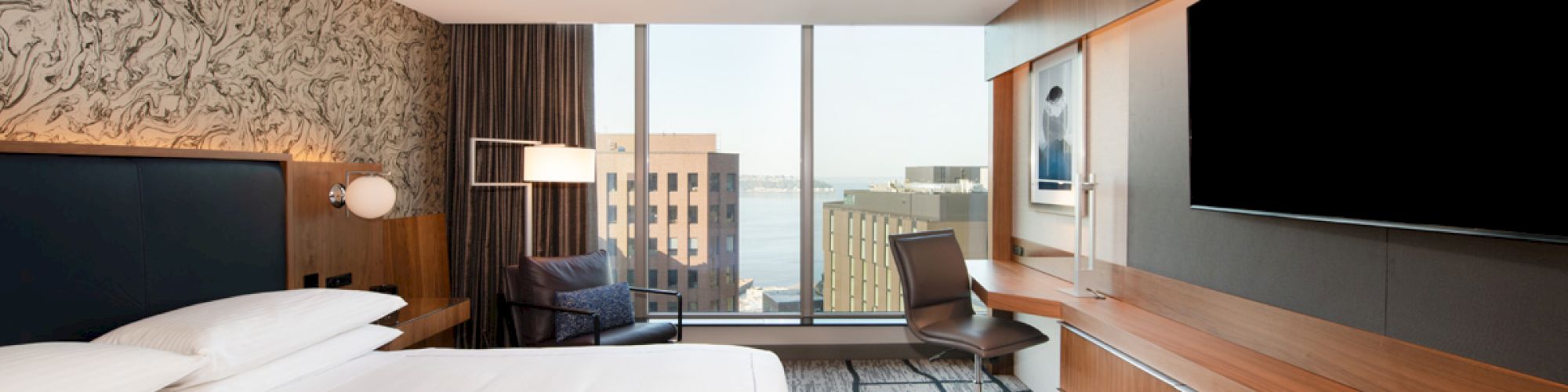 A modern hotel room features a large bed, TV, desk, chair, and a view of buildings through a wide window, with a textured accent wall.
