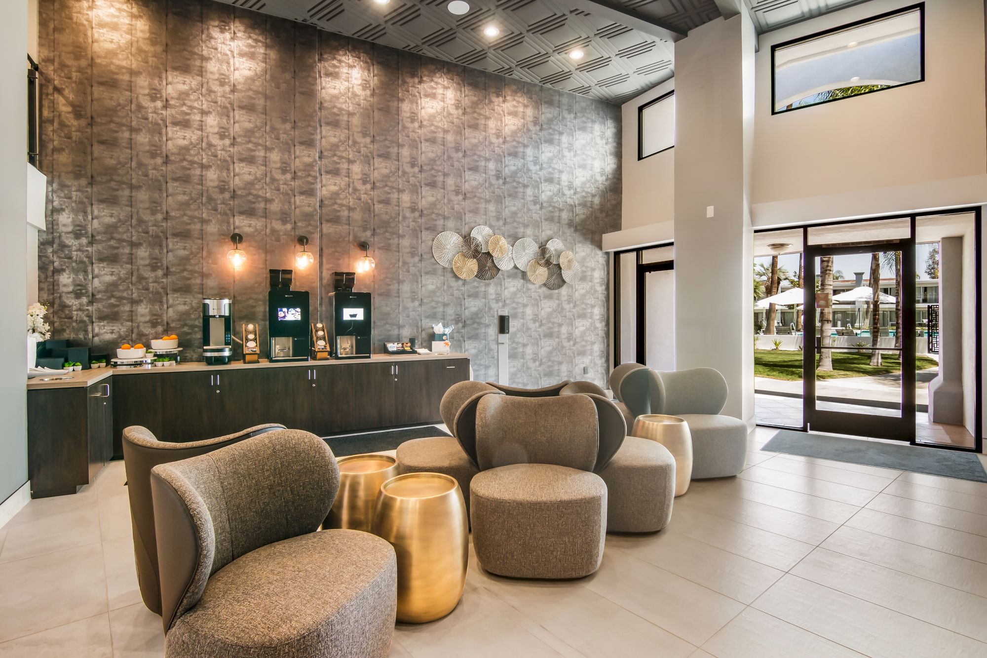 A modern lobby features stylish chairs, golden tables, and a coffee station against a textured wall, with an outdoor view through glass doors.