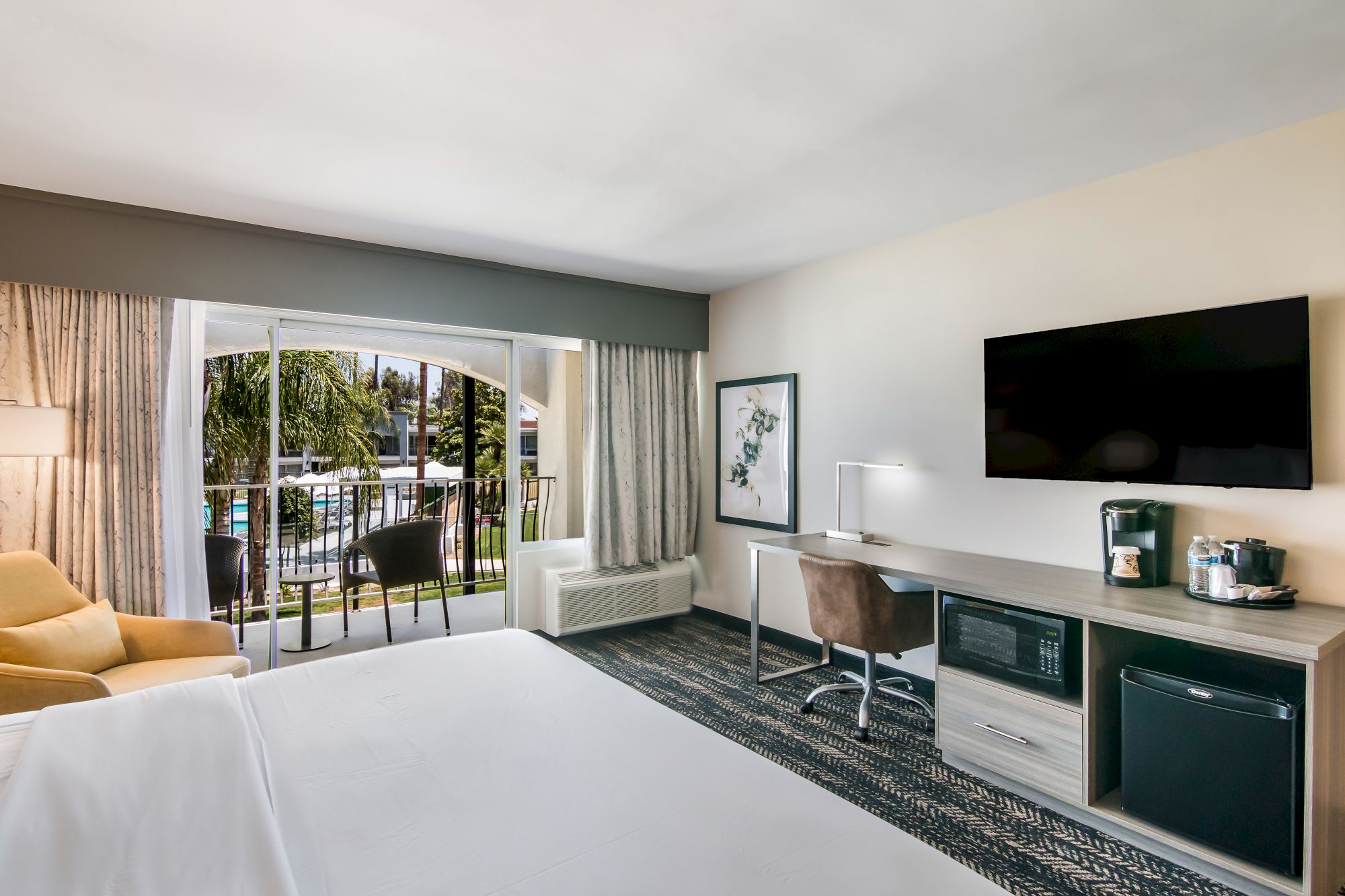 A modern hotel room features a bed, desk, chair, TV, mini-fridge, and coffee maker, with a balcony overlooking a pool area, and ample natural light.